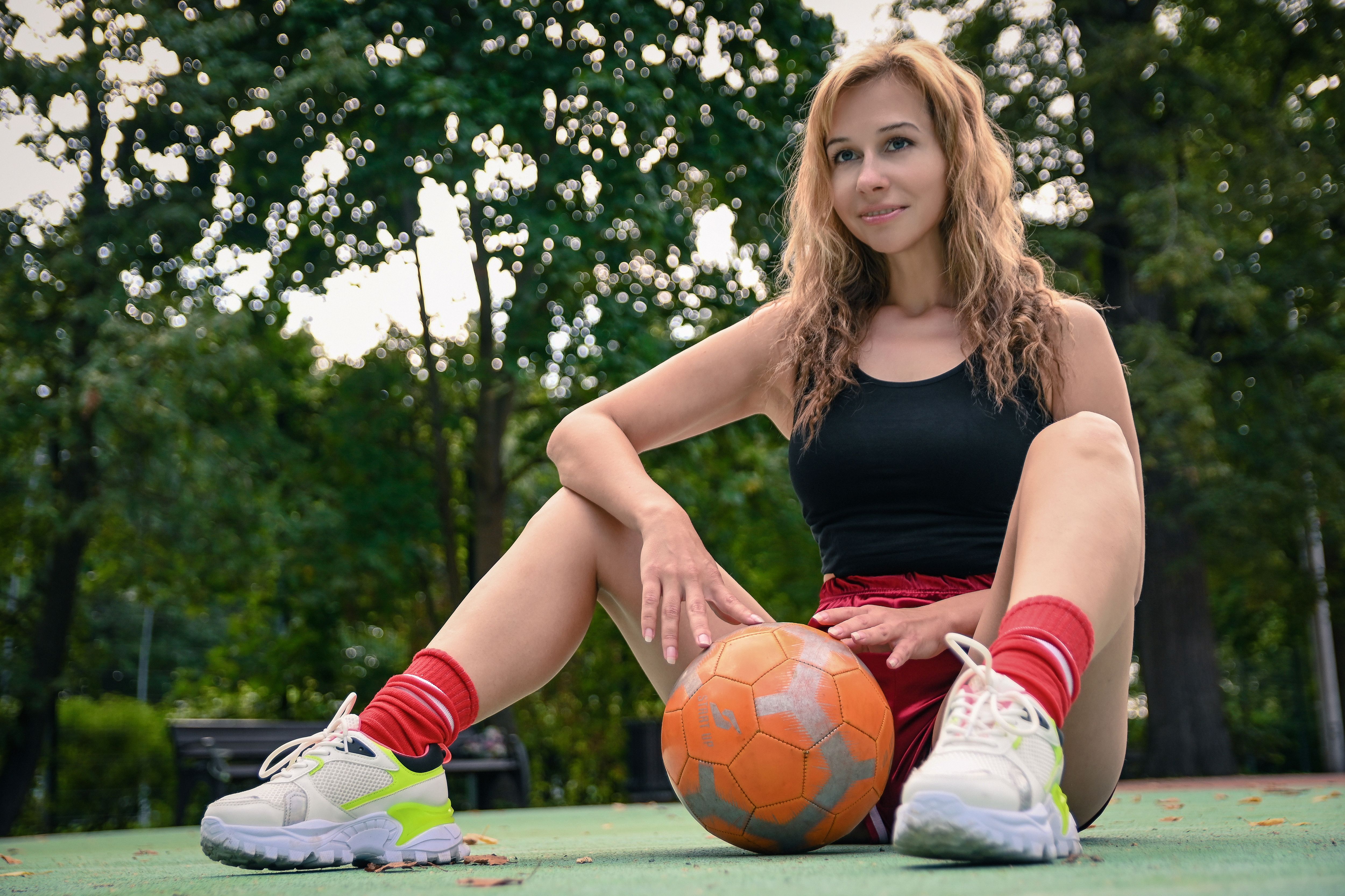 Smiling young woman in sportswear sitting on court with a soccer ball. Outdoor summer portrait.