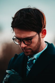 Portrait of a young man with eyeglasses, dressed elegantly, standing outdoors in thoughtful pose.