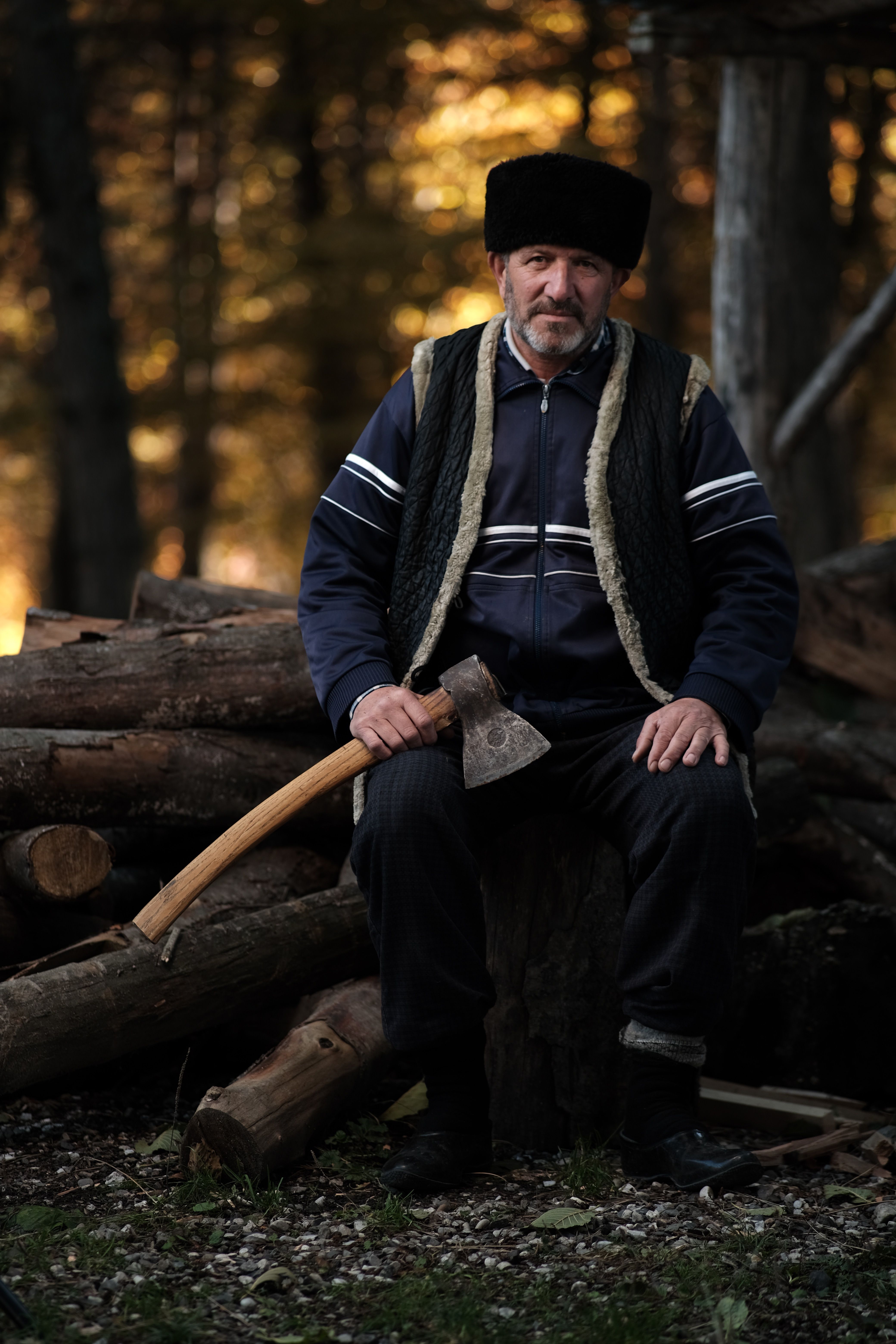 Man sitting on a log pile in a forest with an axe, wearing traditional clothing.