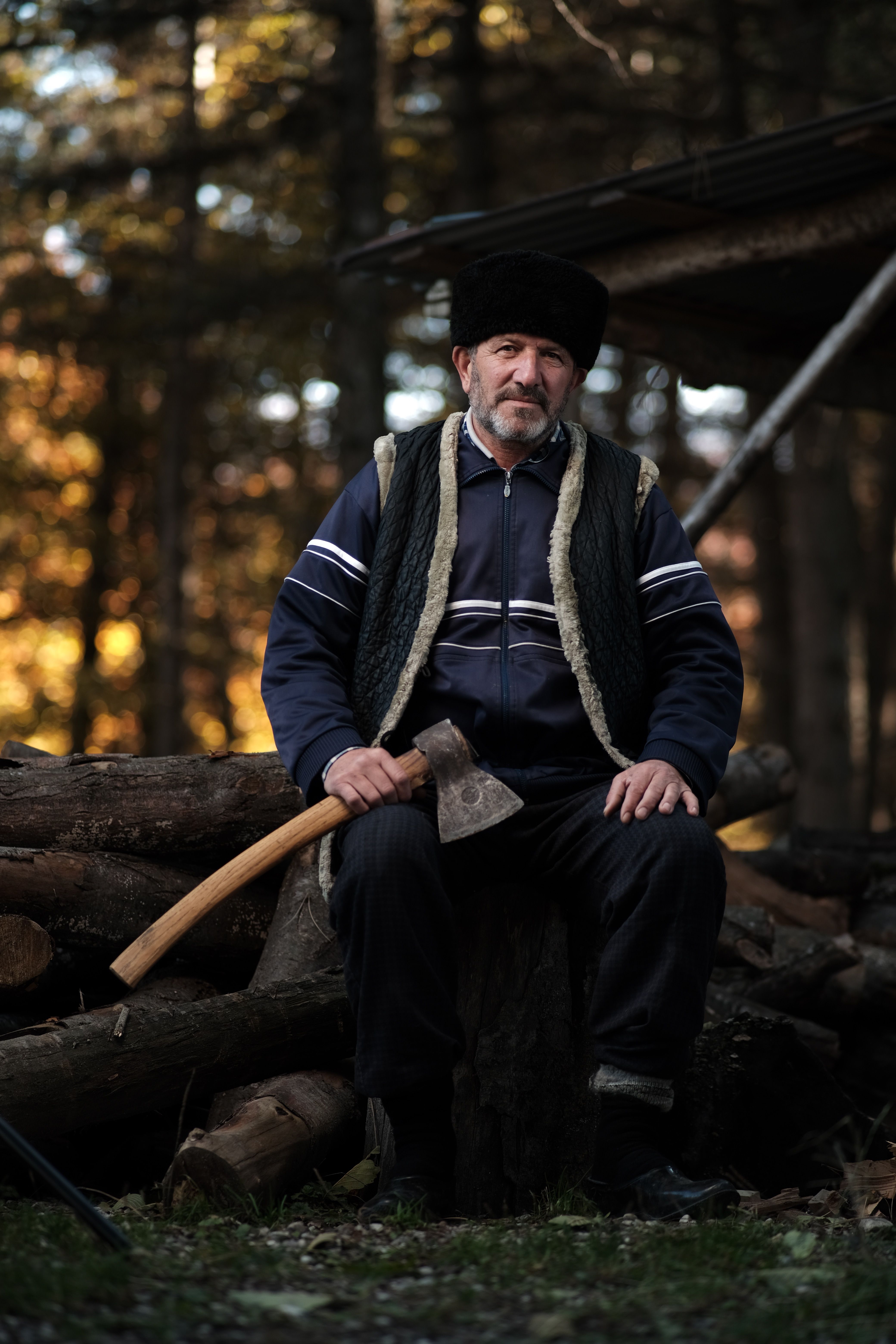 An elderly man with a fur hat and axe seated by wood piles in a rural village setting.