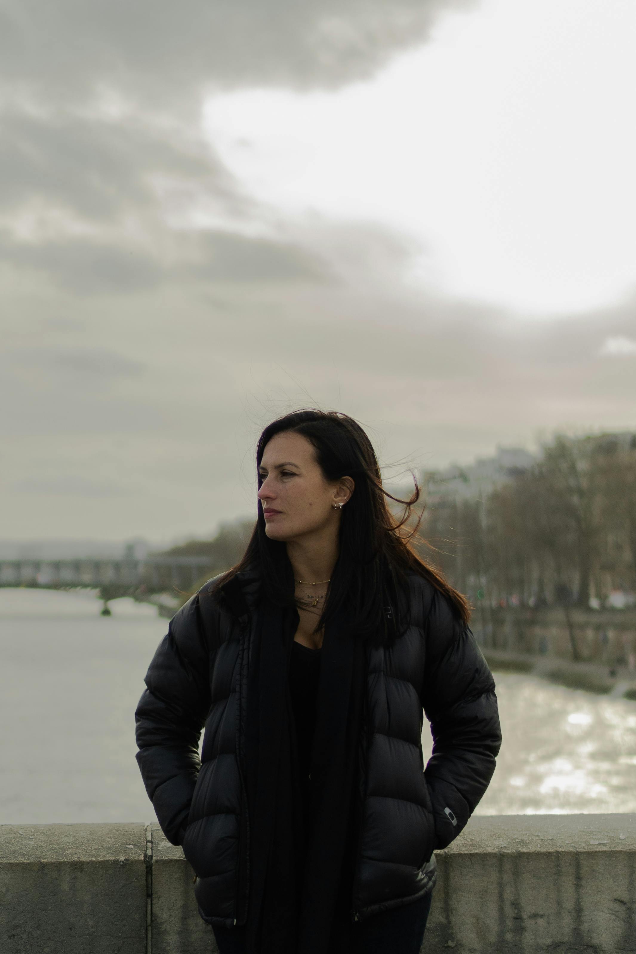 Portrait of a woman in a black jacket standing by a river with cityscape in the background.
