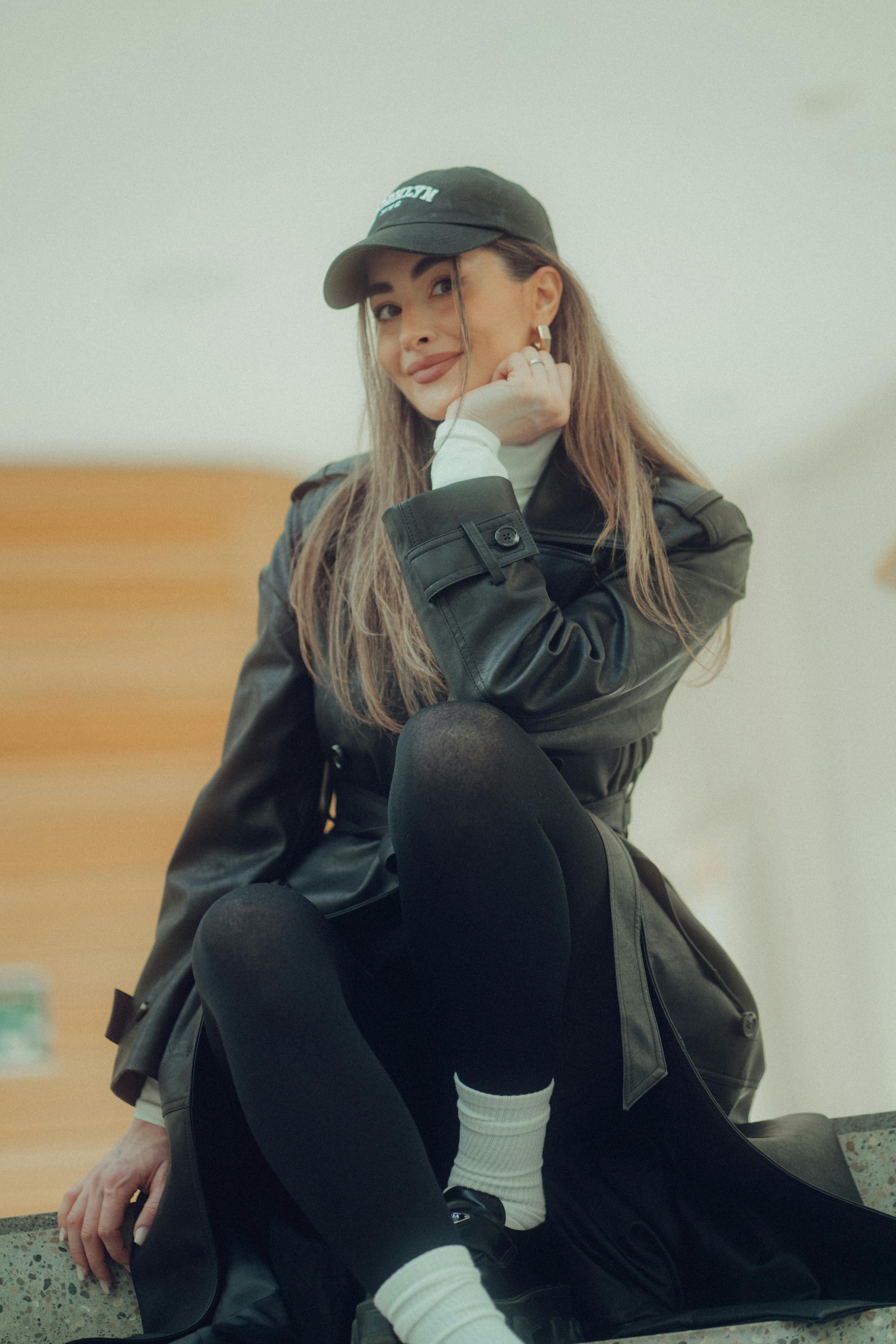 Portrait of a fashionable young woman sitting indoors wearing a black overcoat and baseball cap.