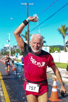 Elderly man pouring water over head while running in sunny urban marathon.
