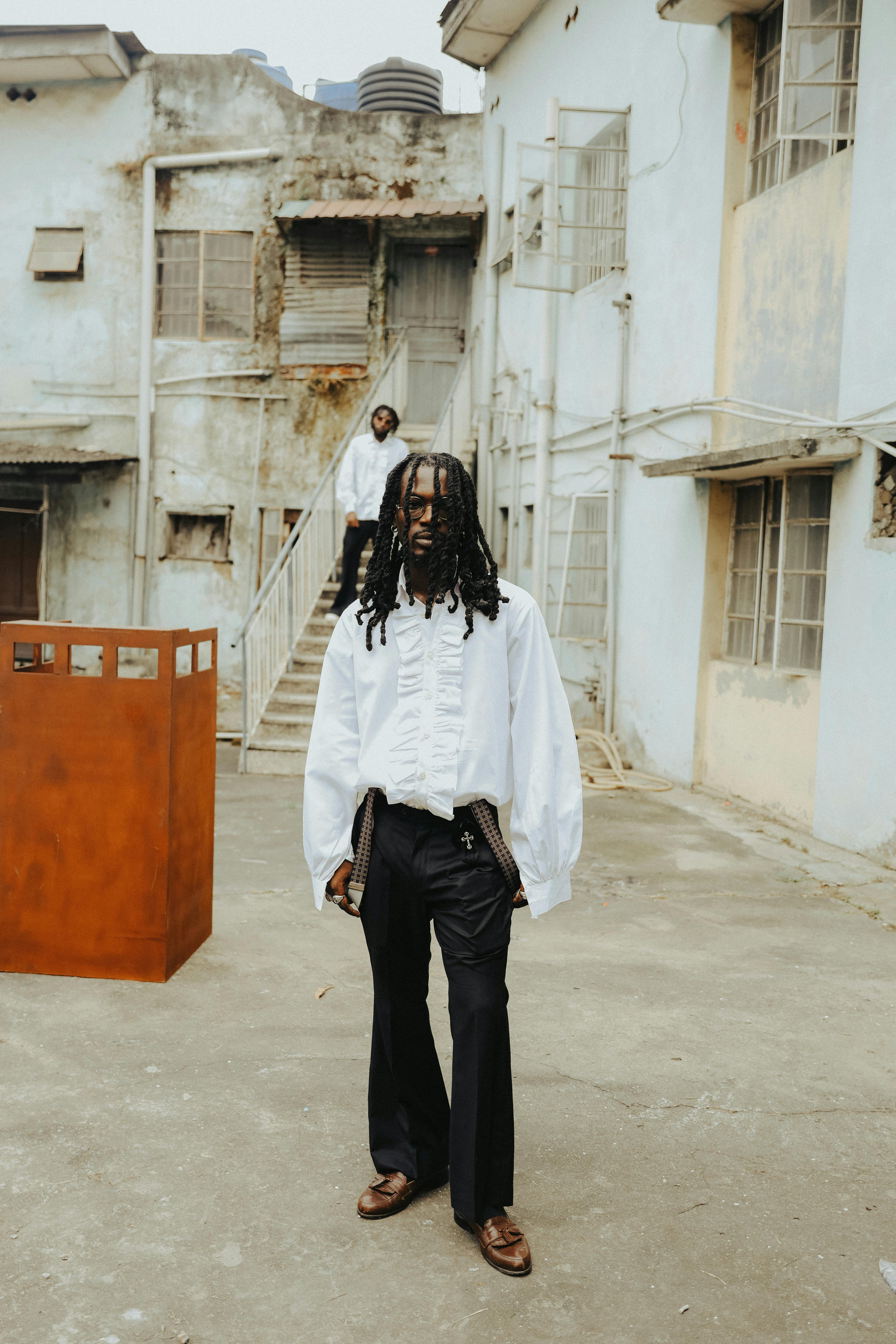 Stylish man with braided hair in white shirt stands in front of rustic urban building.
