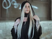 一位戴着黑色兜帽、留着金色长发的优雅女子在室内摆着姿势