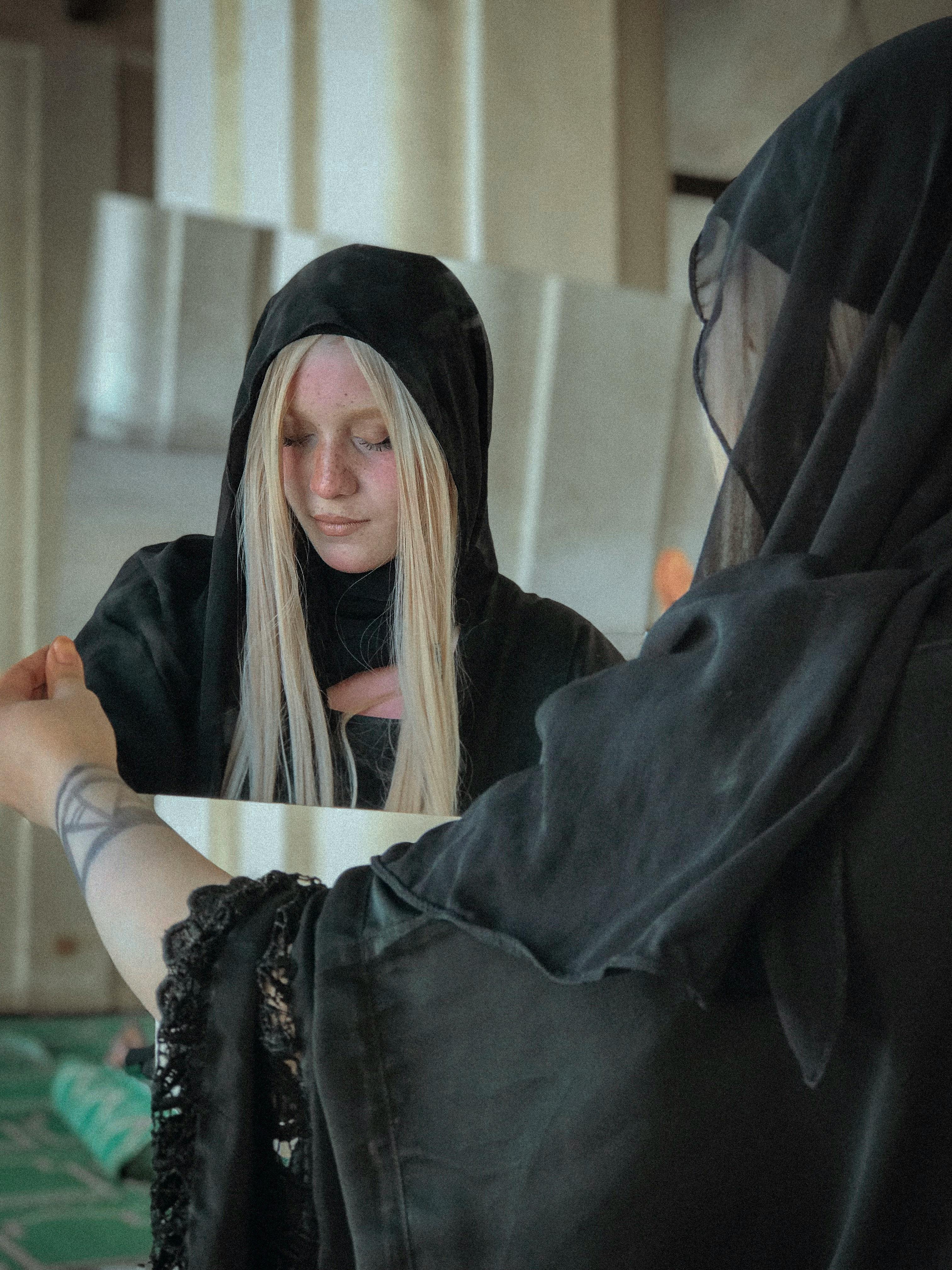 一名戴着黑色兜帽、留着金色长发的女子凝视着镜中的自己