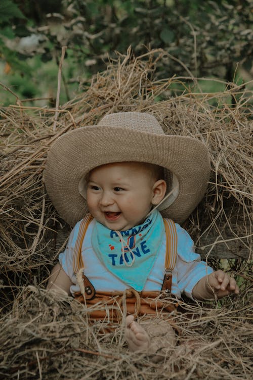 A baby wearing a cowboy hat and bandana sitting in hay