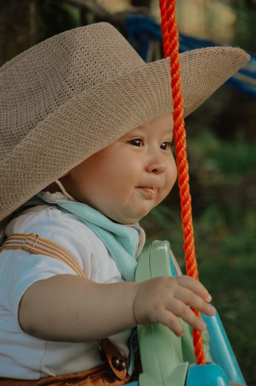 Free Charming baby wearing a straw hat, sitting on a swing in a garden setting, radiating joy and innocence. Stock Photo