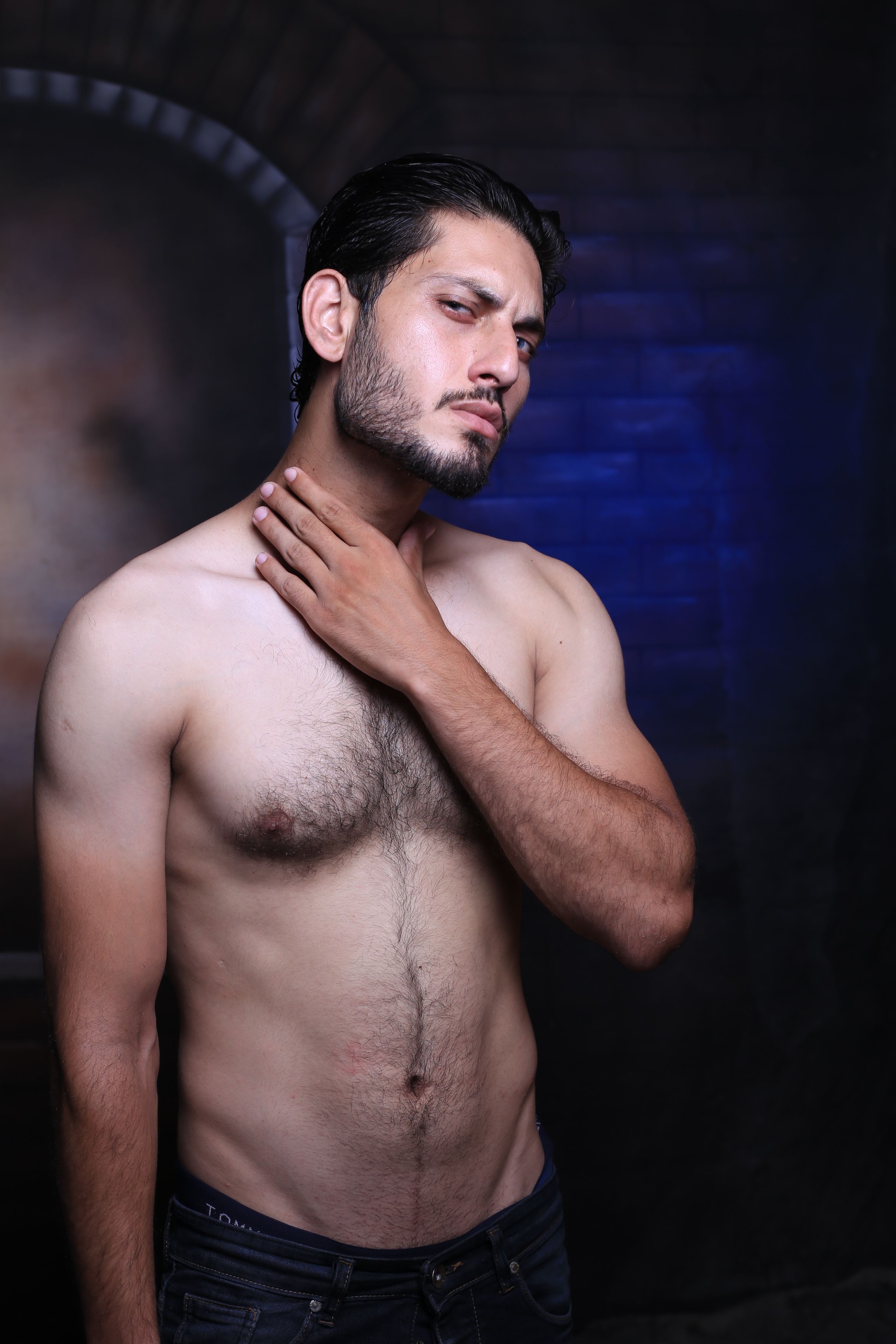 Fashion portrait of a young man with black hair touching neck, photographed indoors.