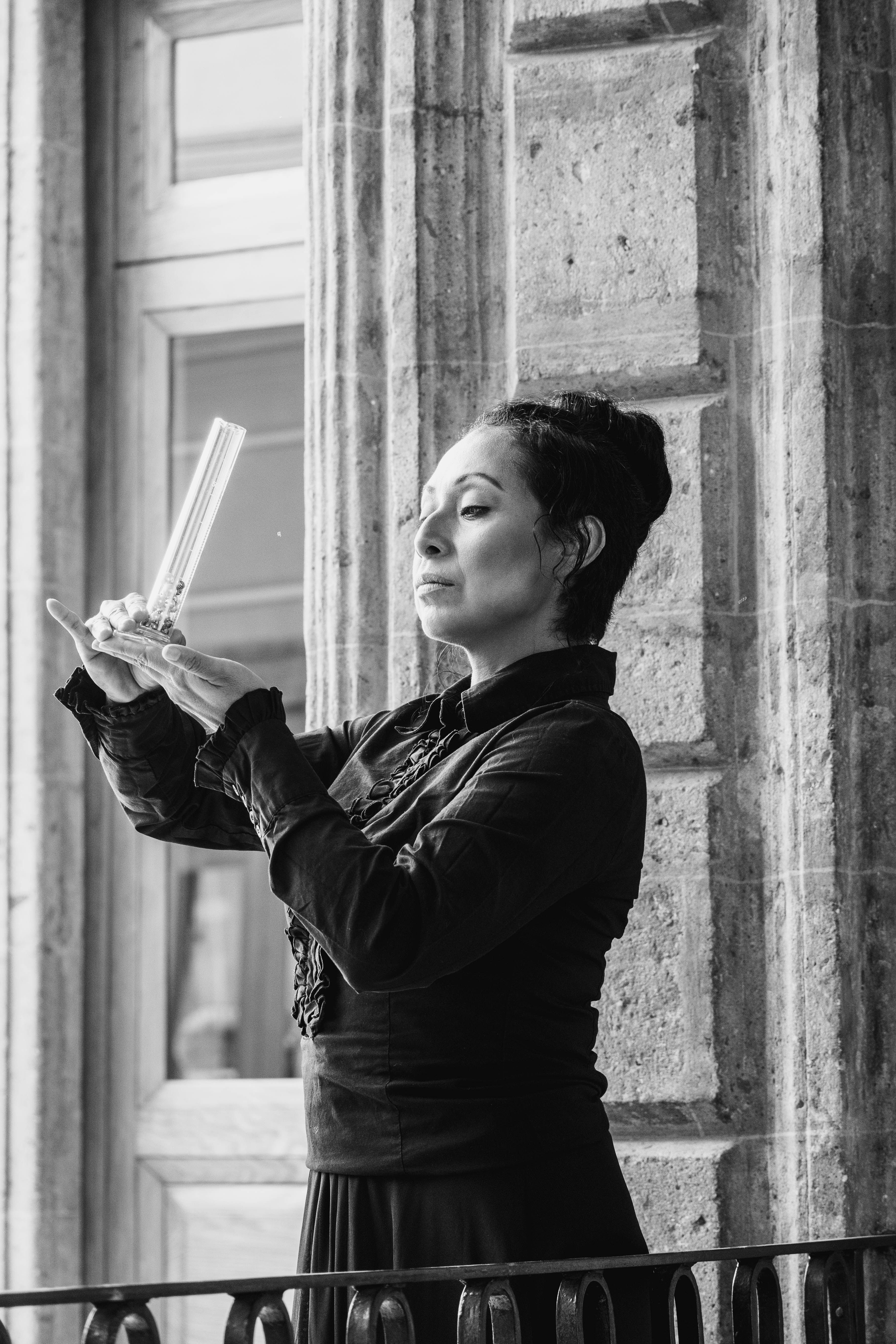 Black and white portrait of a woman holding a glass cylinder at a balcony in Mexico City, showcasing classic fashion.