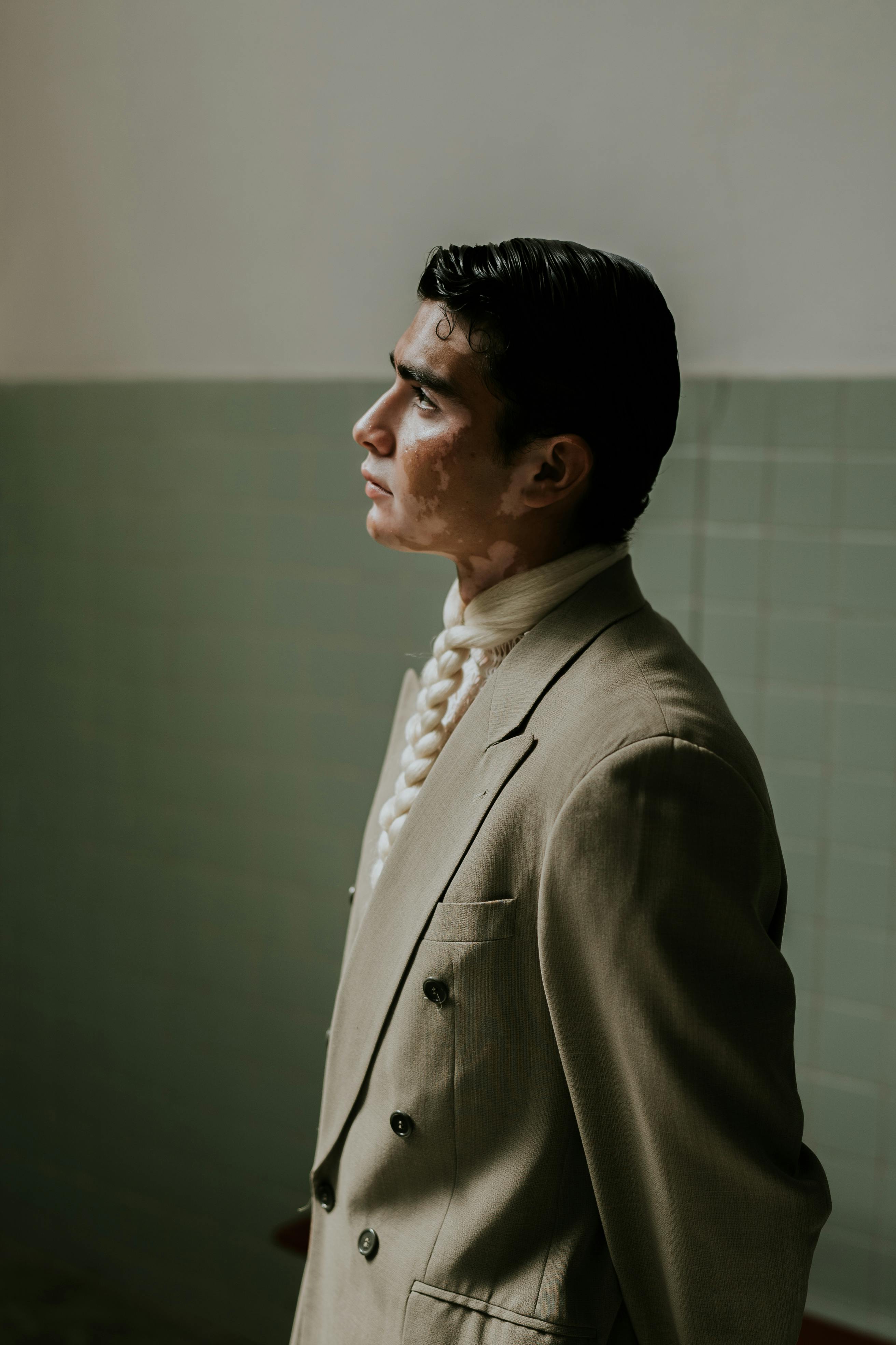 Side profile of a man in an olive blazer and braided tie posing indoors.