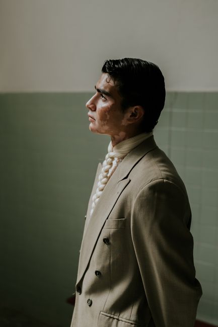 Side profile of a man in an olive blazer and braided tie posing indoors.