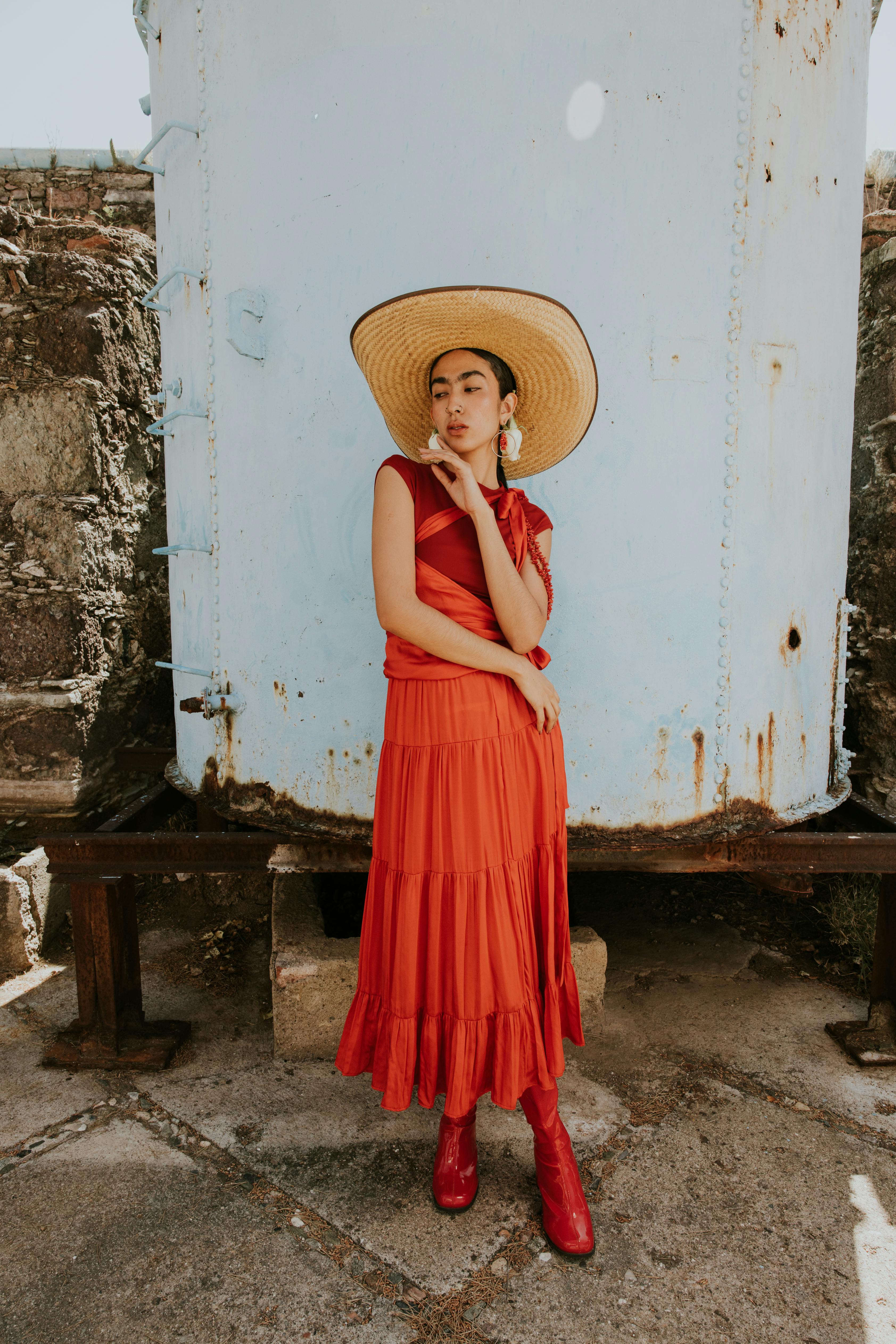 Elegant woman in a red dress and straw hat posing outdoors, capturing urban fashion.