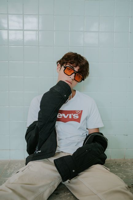Fashionable young man with sunglasses poses indoors against a tiled wall.