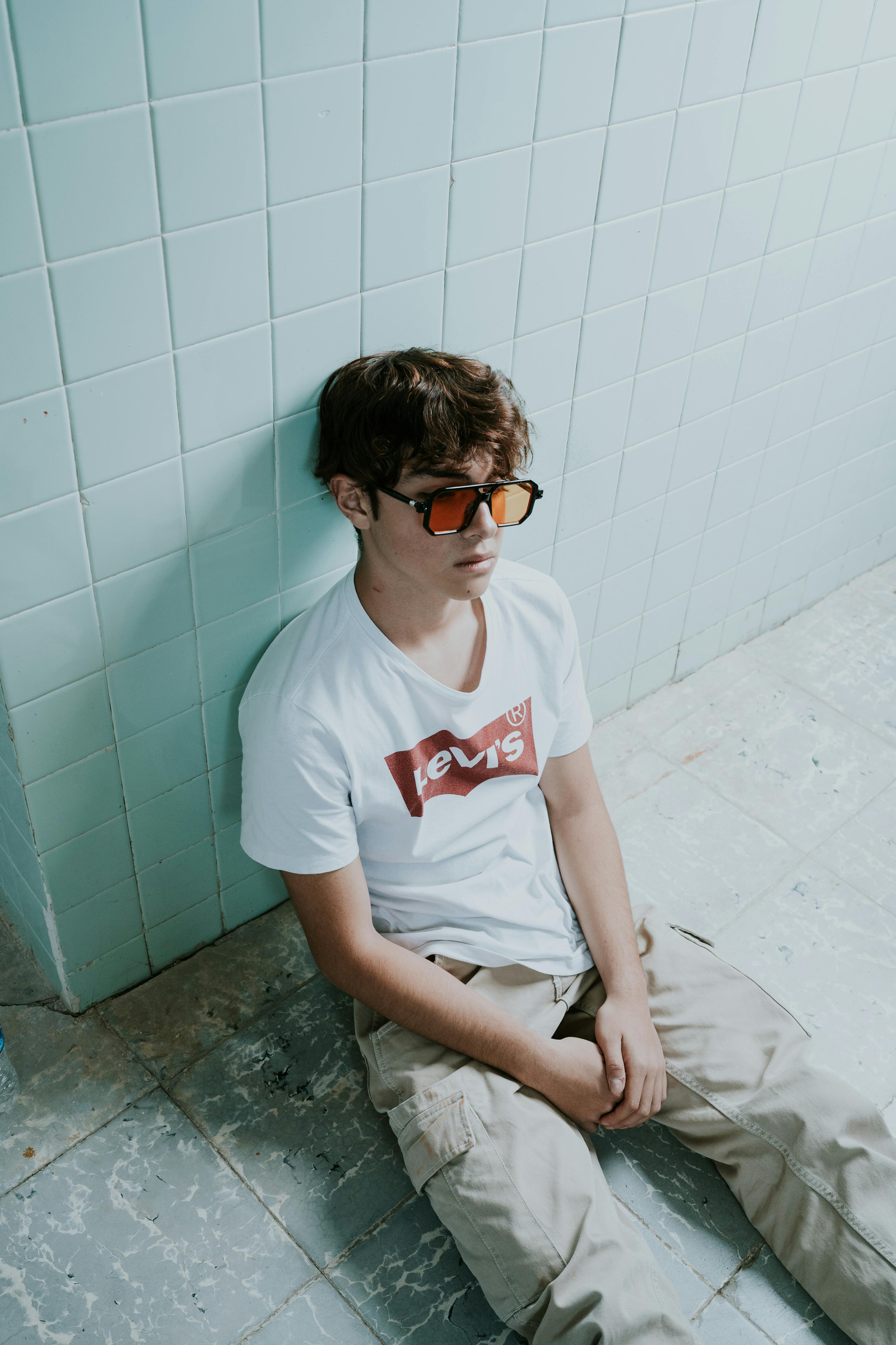 A fashionable young man wearing sunglasses and a white T-shirt sits against a tiled wall.
