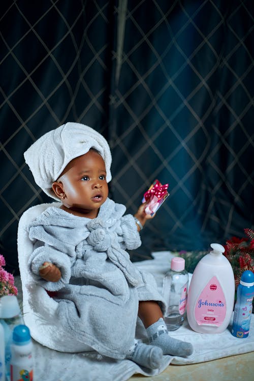 A baby in a bathrobe sitting on a chair with a bottle of lotion