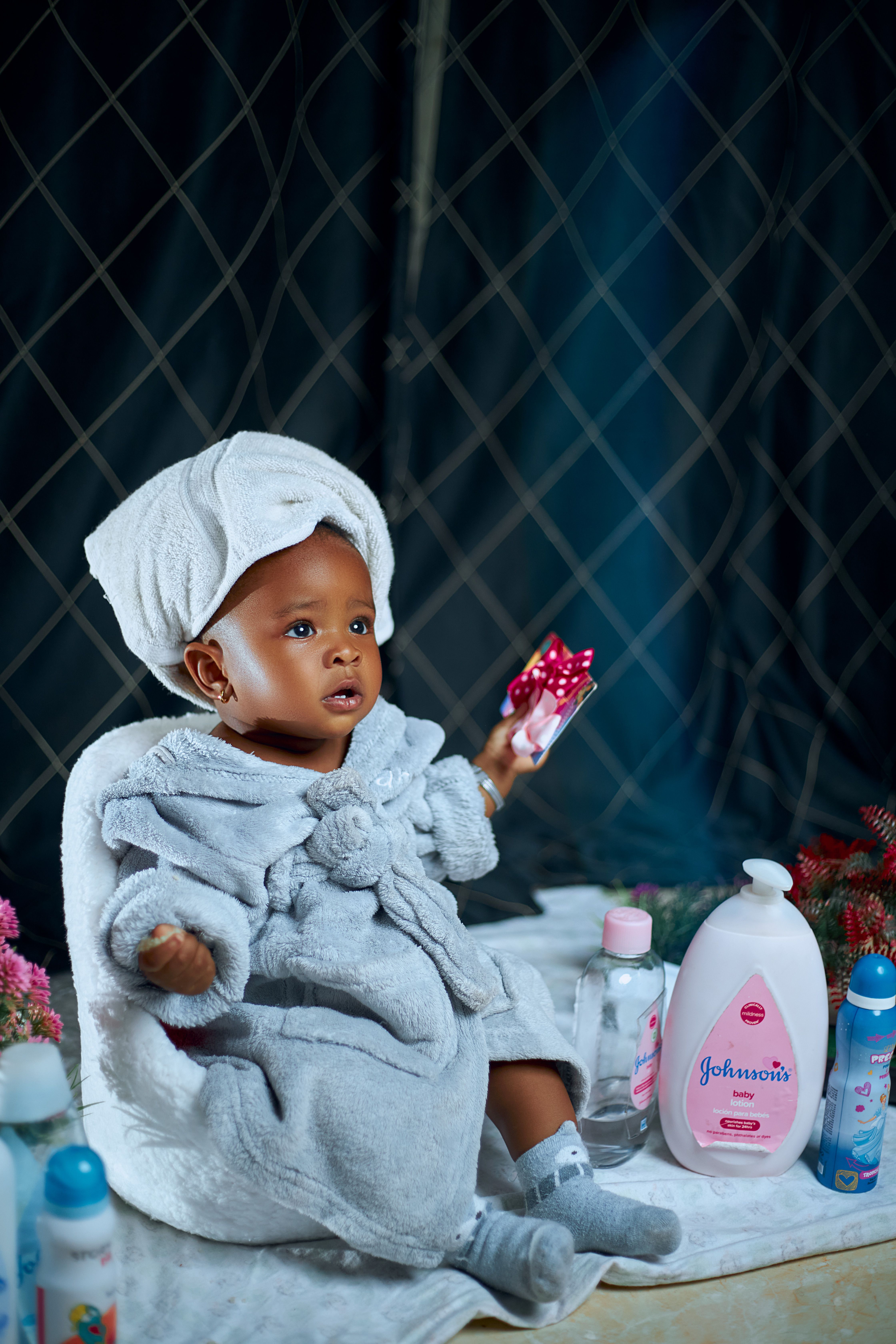 Free A cute toddler in a bathrobe holding a toy, surrounded by baby toiletries, sitting indoors. Stock Photo