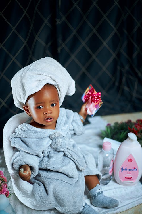 A baby in a bathrobe sitting on a toilet with a bottle of lotion