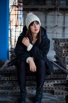 A young woman in a white hat and black jacket poses stylishly at a construction site.