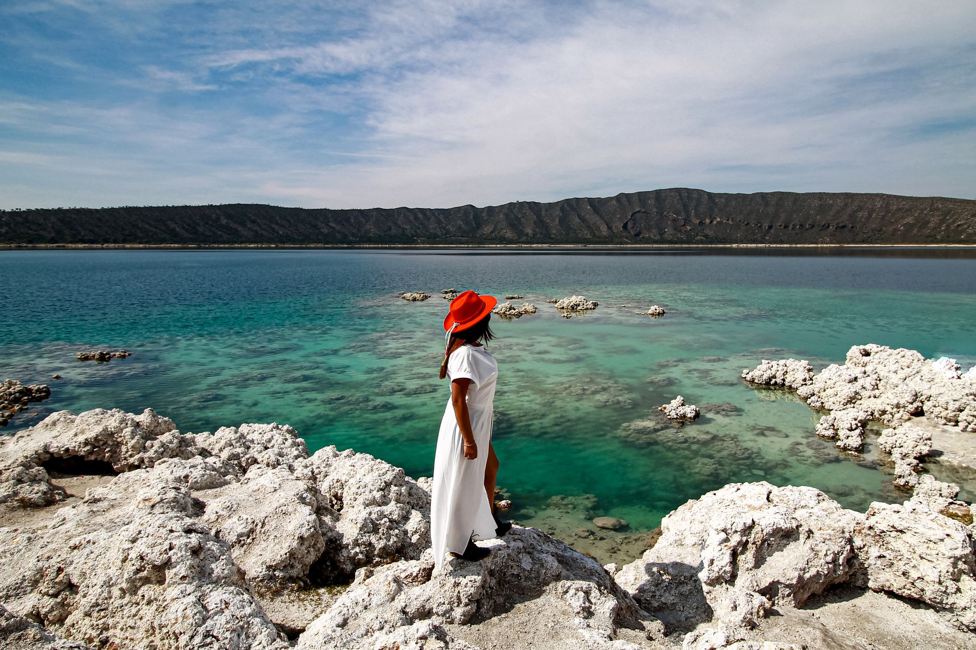 A woman gazes over turquoise waters at Laguna Alchichica, Mexico, wearing a red hat and white dress.