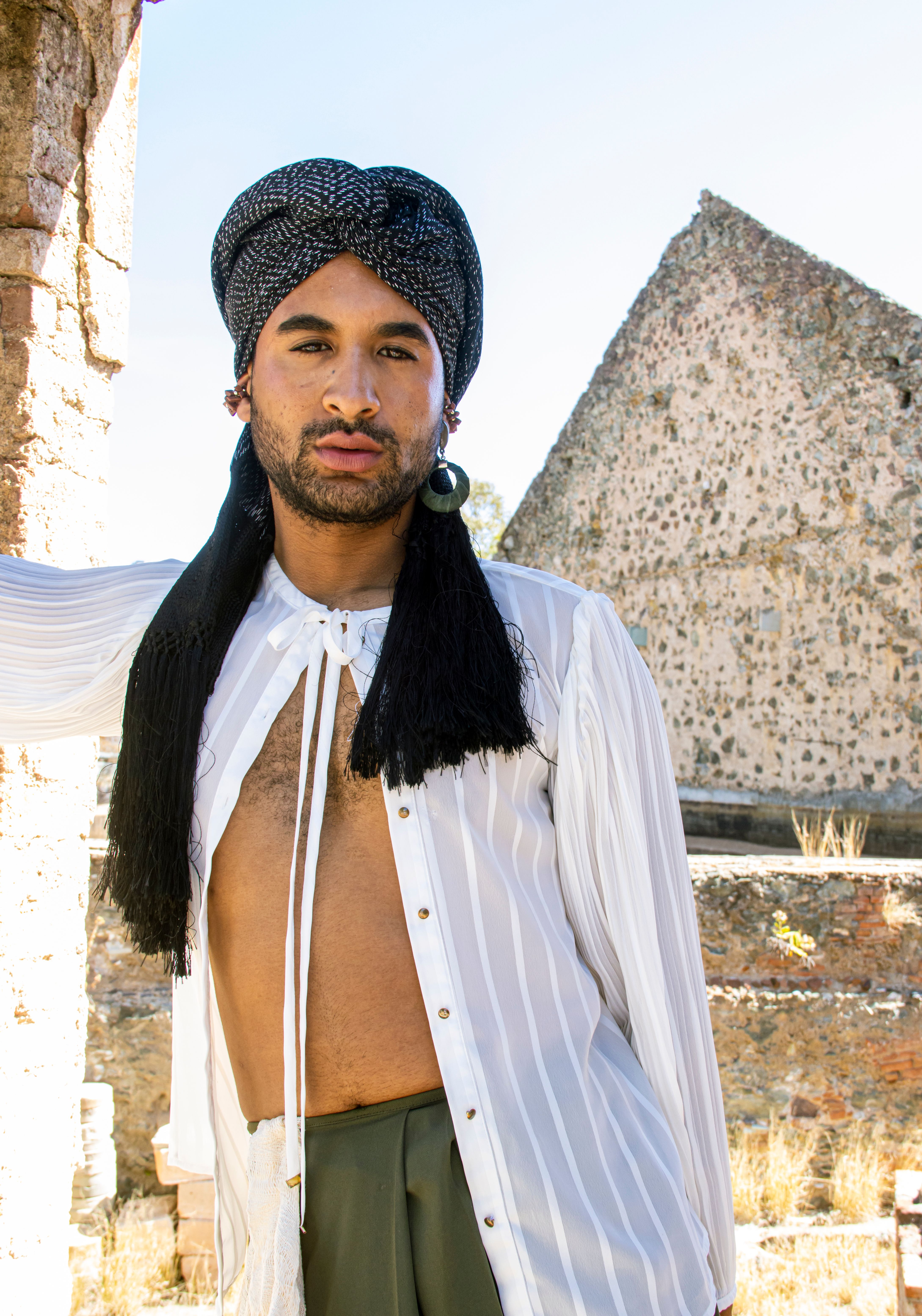 Fashion portrait of an adult man wearing traditional headwear and a white shirt, set outdoors in daylight.