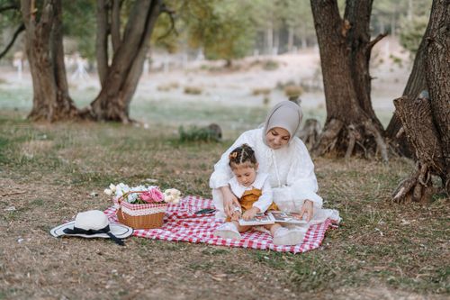 Free A mother and daughter enjoy a peaceful picnic under trees, reading a book together. Stock Photo