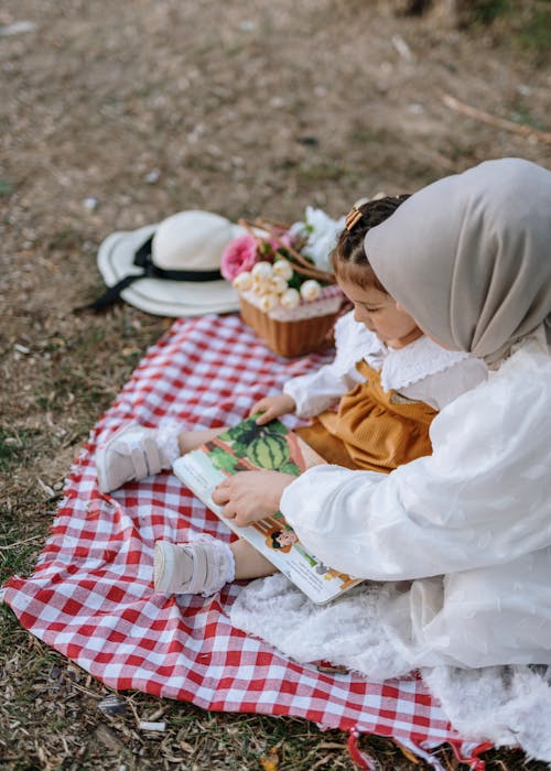 A woman and child sitting on a blanket reading a book