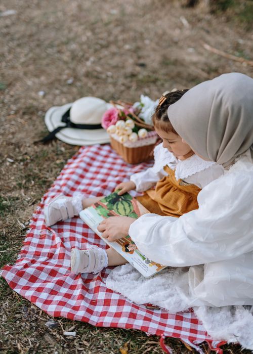 Free A woman in a hijab reading a book with her daughter on a picnic blanket outdoors. Stock Photo