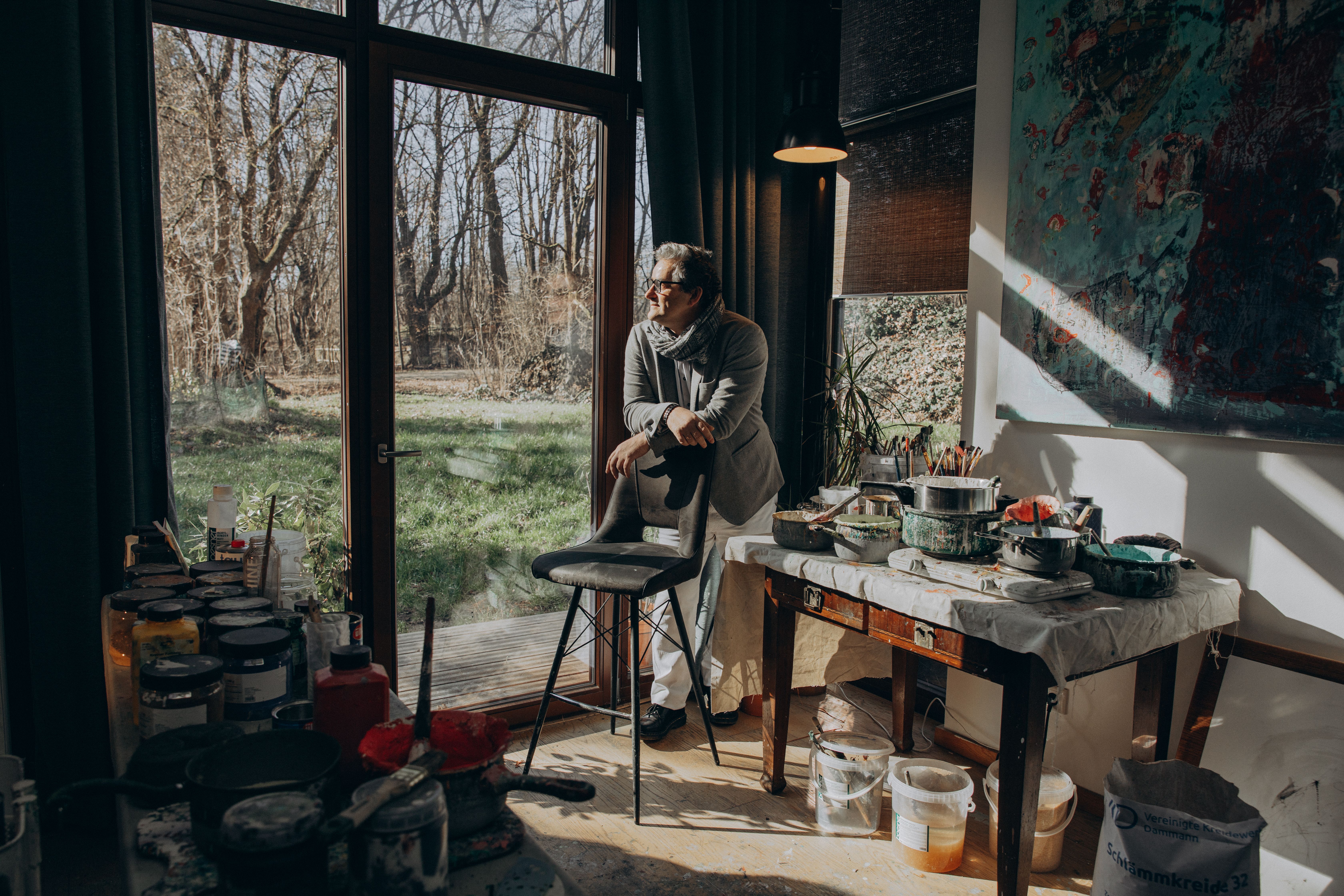 A senior male artist gazes outside the window in his sunlit studio filled with painting supplies.