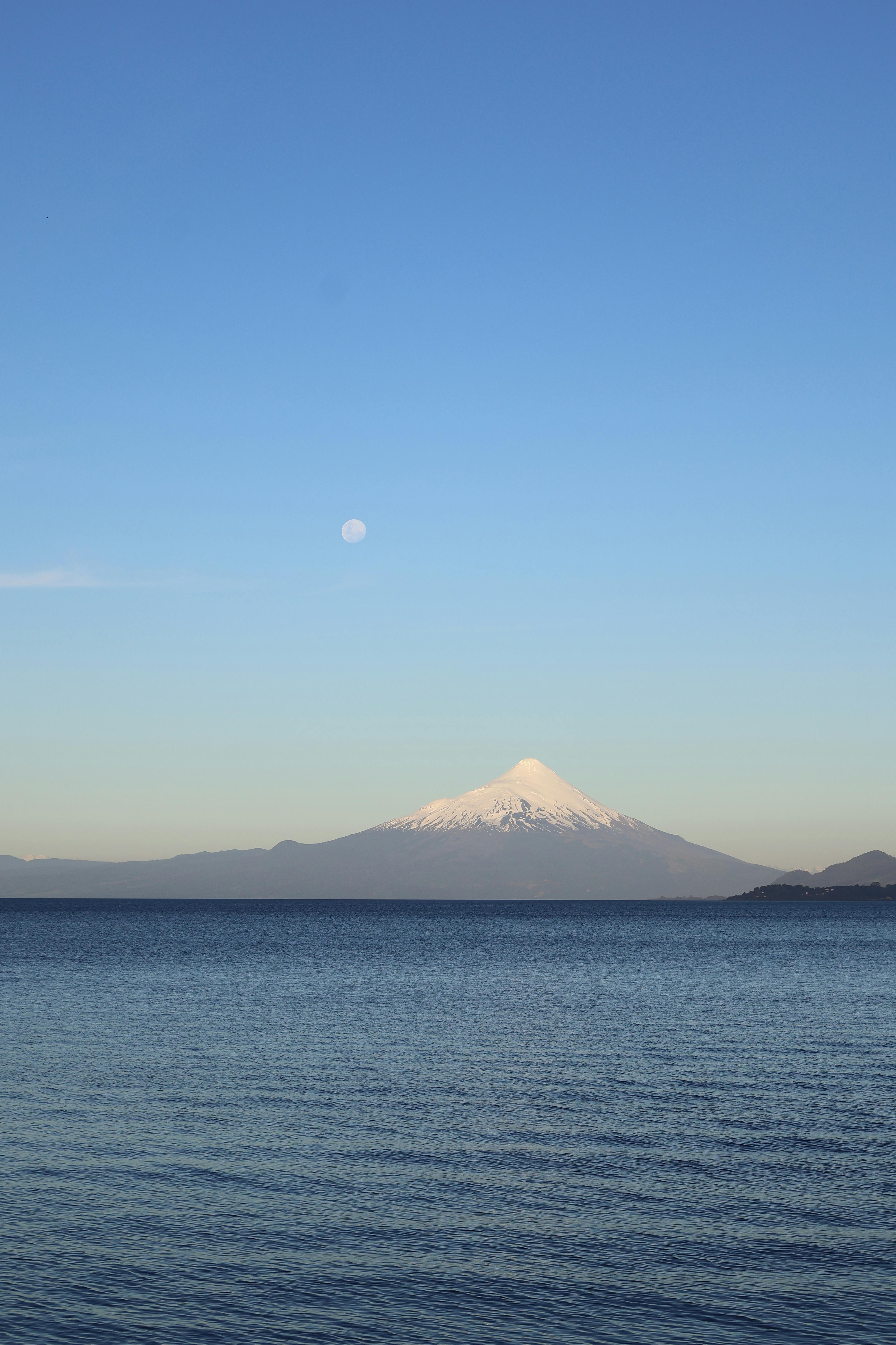 Serene view of a snow-capped mountain with a full moon above and tranquil water below.