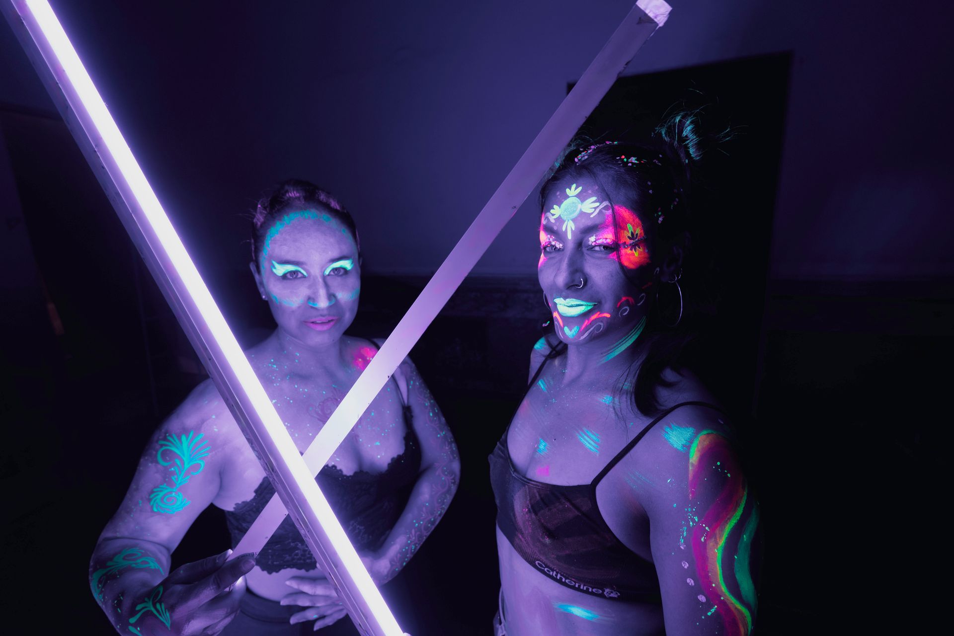 Two women in vibrant neon body paint posing under UV lights in a dark studio setting.