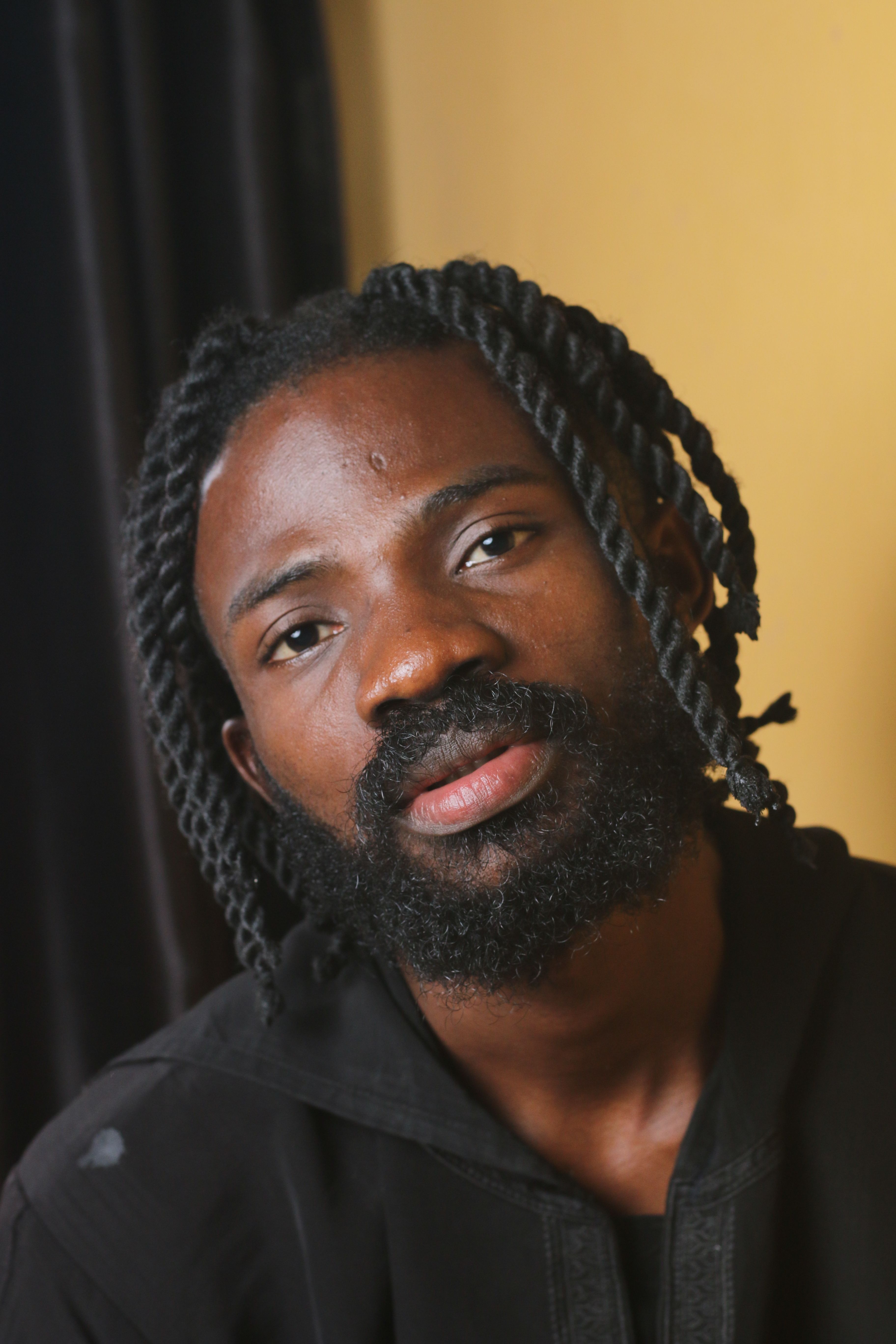 Close-up portrait of a man with a beard and twisted hairstyle against a neutral background.