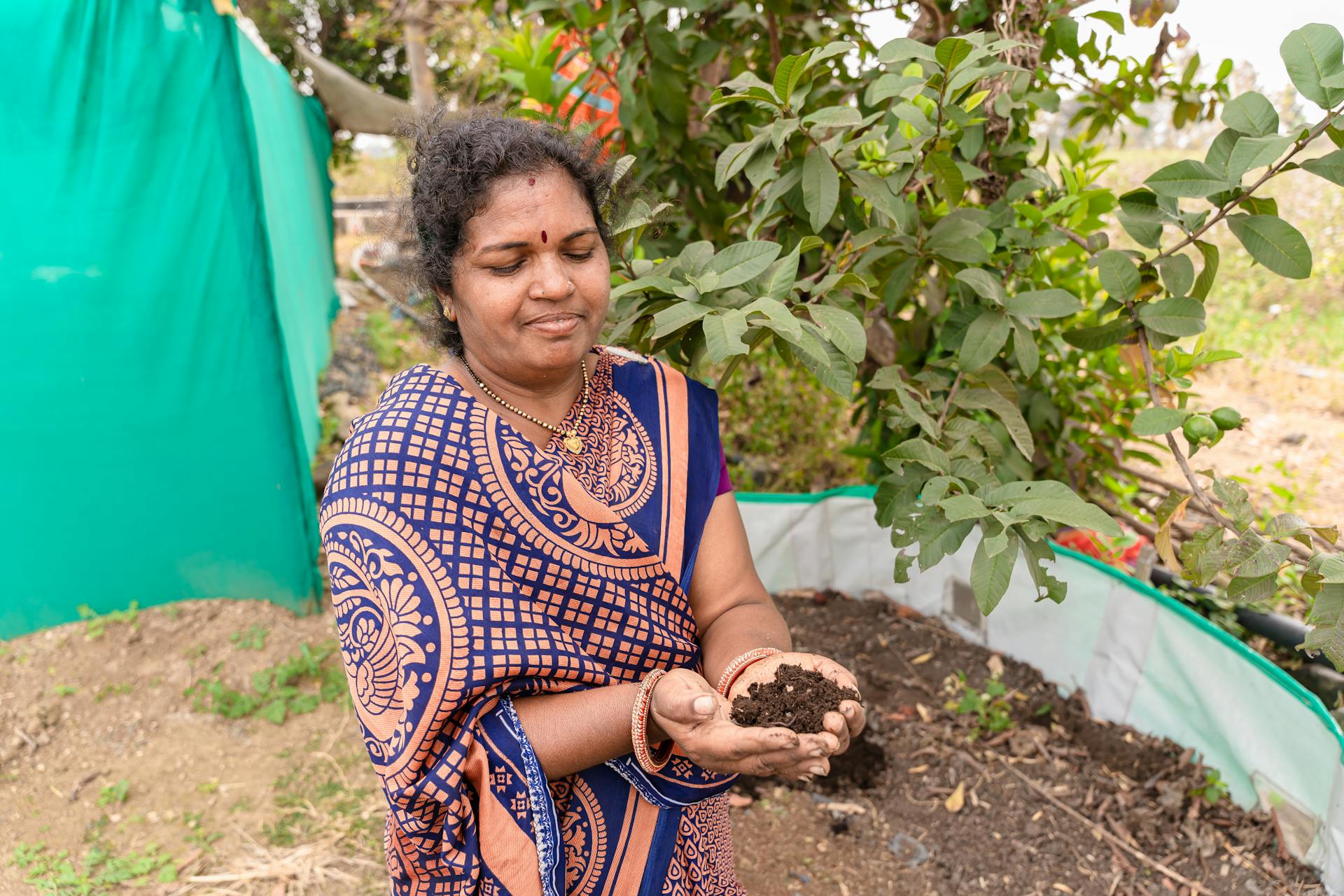 South Asian woman in traditional attire holding soil in a garden setting, promoting sustainable agriculture.