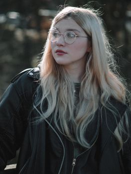 Teenager wearing a leather jacket and glasses, posing thoughtfully outdoors.