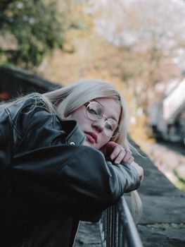 A young woman in a leather jacket with eyeglasses leans on a railing outdoors, embodying style and contemplation.