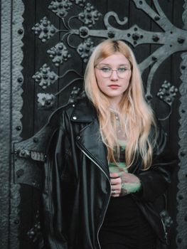 Elegant young woman with glasses posing in a leather jacket against an ornate gate backdrop.