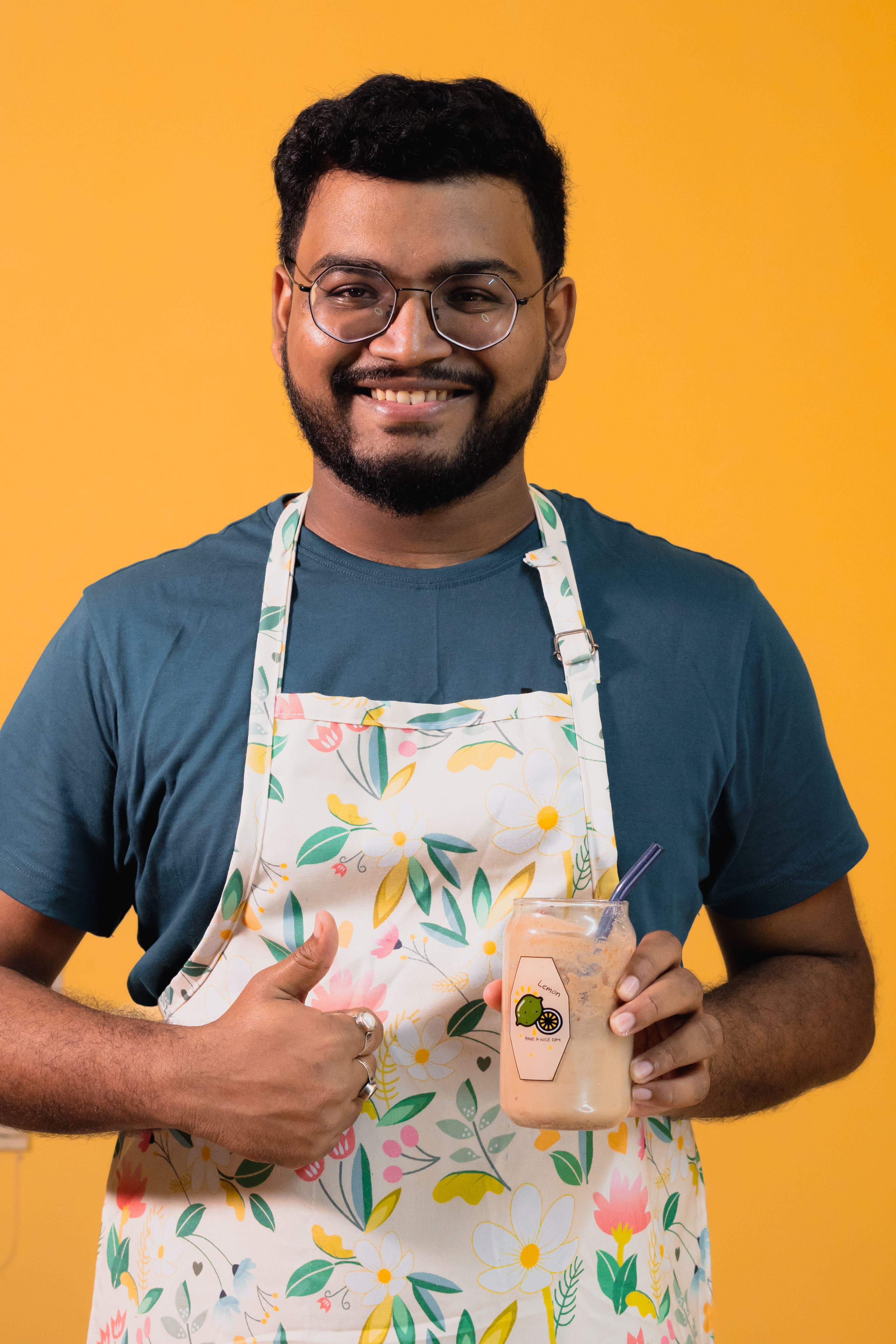 Portrait of a smiling man in a floral apron holding juice against a vibrant yellow background.