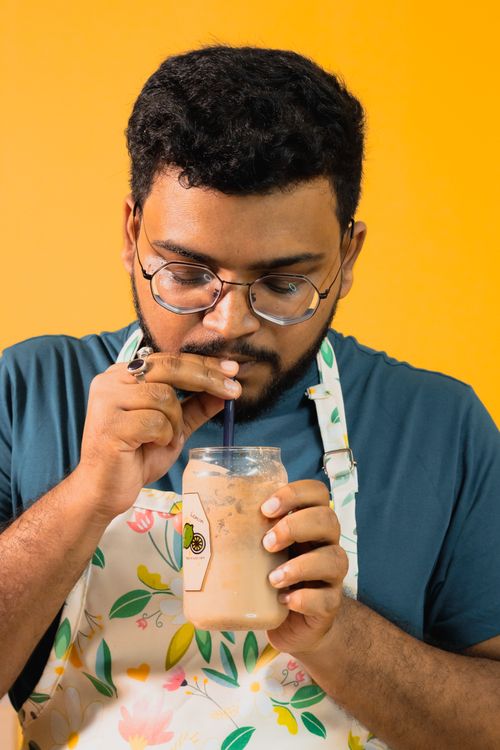 Free Adult male enjoying an iced coffee drink through a straw in a floral apron, set against a vibrant yellow background. Stock Photo