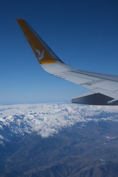 Aerial view of airplane wing flying over majestic snowy mountains with clear blue sky.