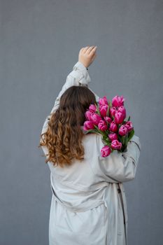 Back view of woman with long hair holding pink tulips against gray background, raising arm.