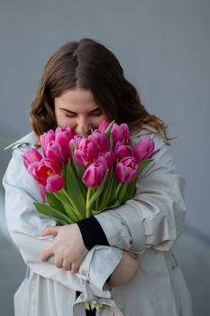 A woman in a trench coat lovingly embraces a bouquet of vibrant pink tulips in a studio setting.