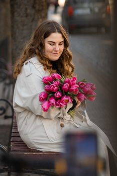 Elegant woman in a trench coat sitting with a bouquet of vibrant pink tulips.