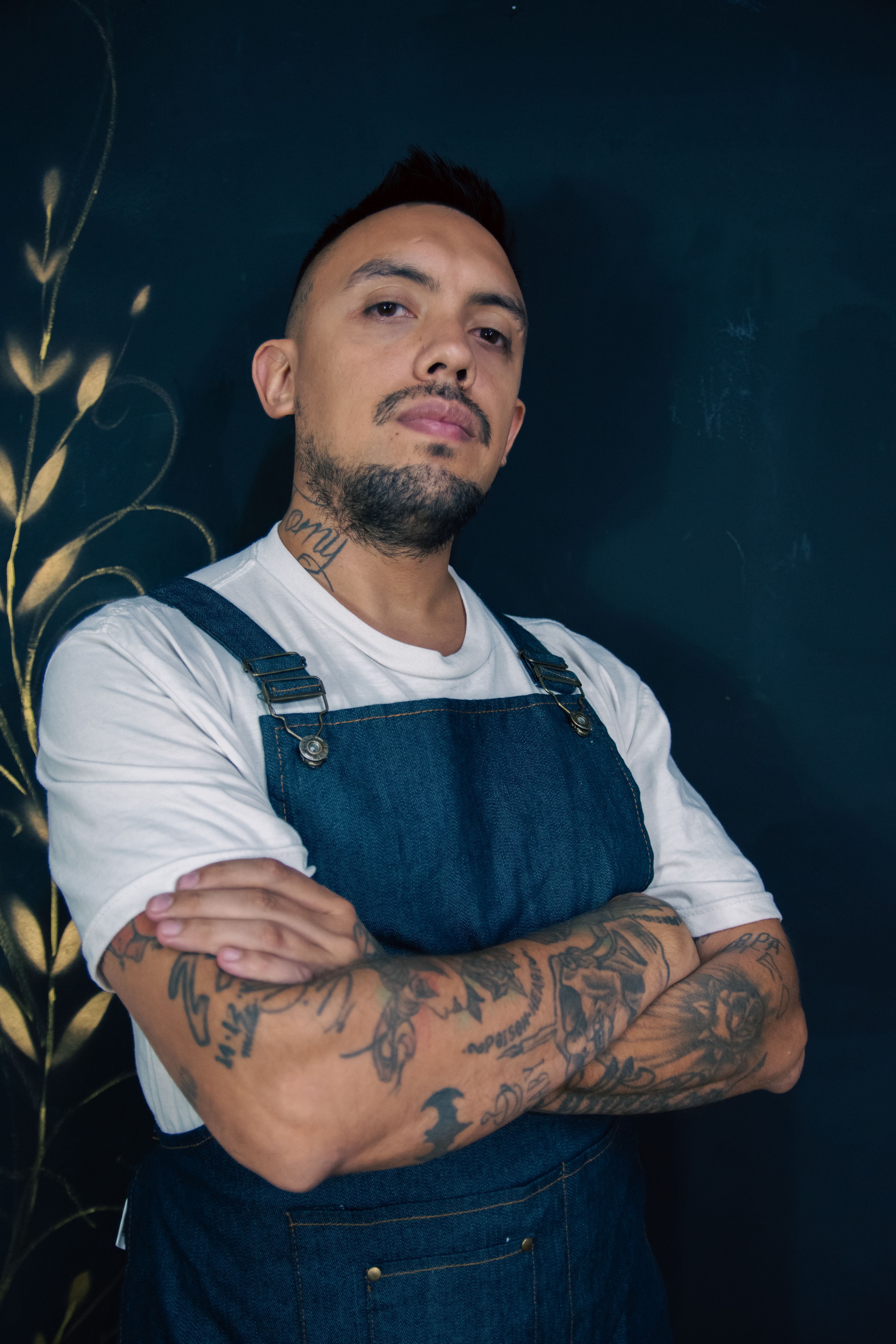 Confident tattooed man posing with arms crossed in a denim apron, showcasing tattoos and style.