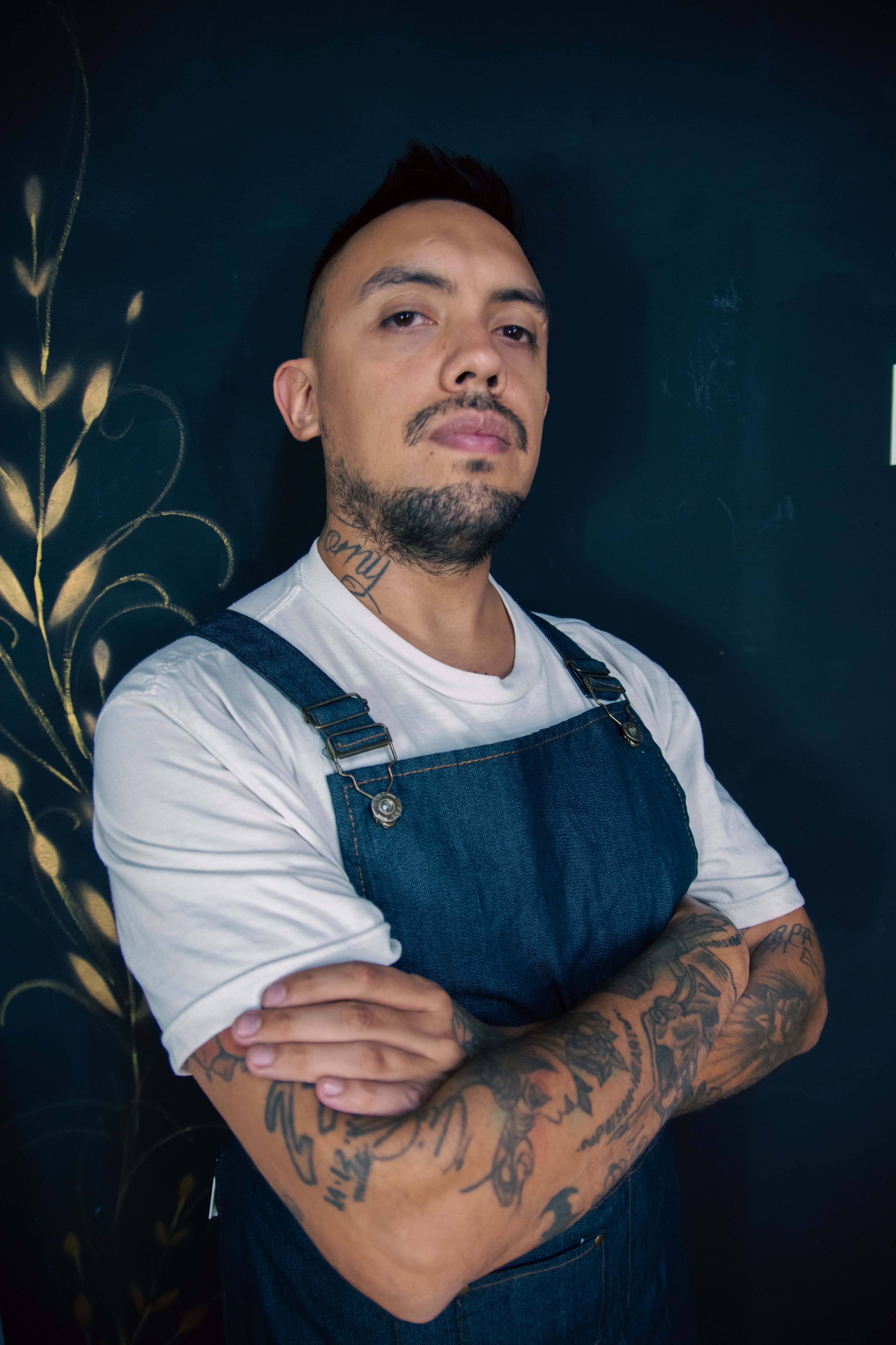 Tattooed man standing confidently with crossed arms in denim overalls against a dark background.