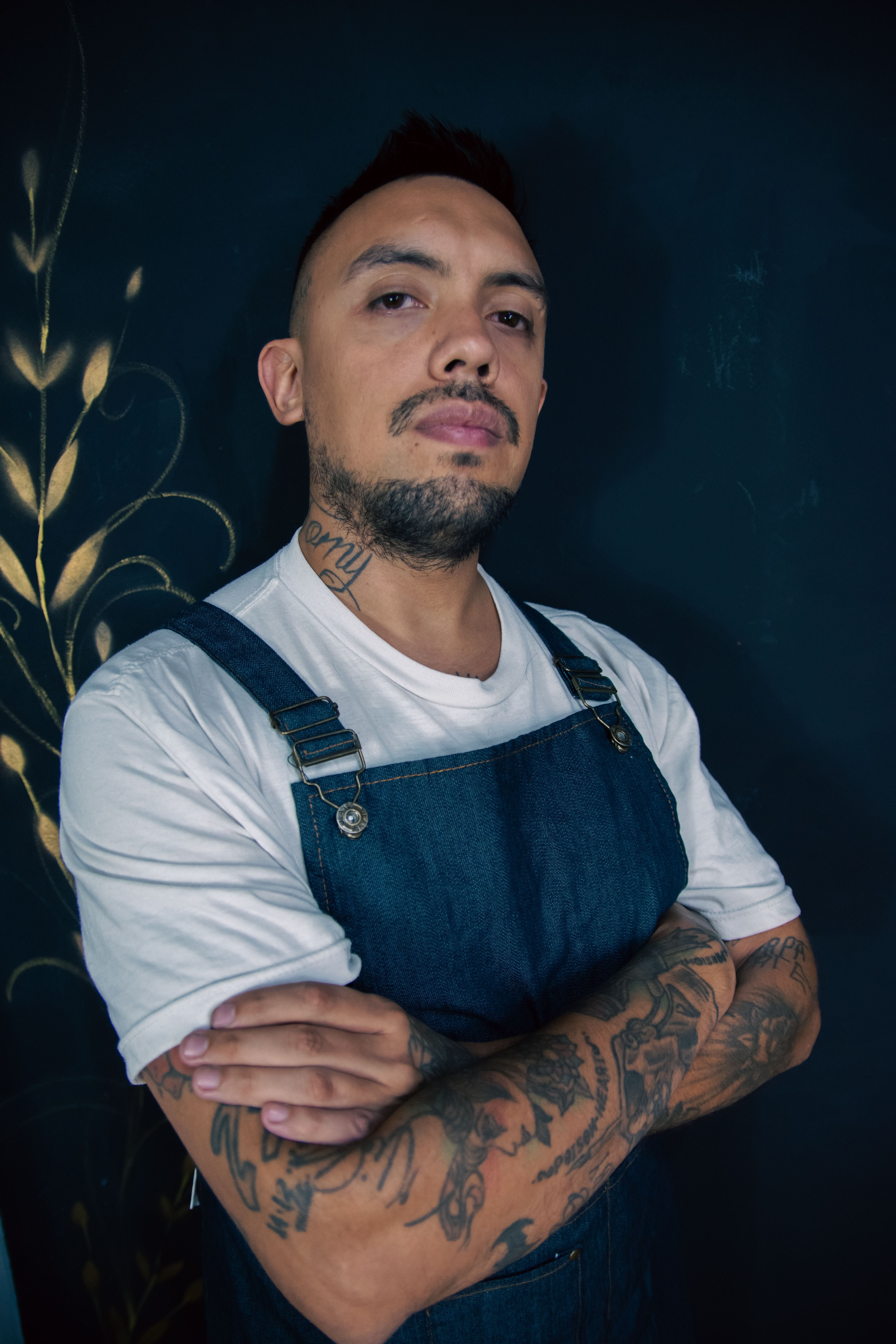 Confident tattooed man in denim apron with arms crossed, indoors portrait.