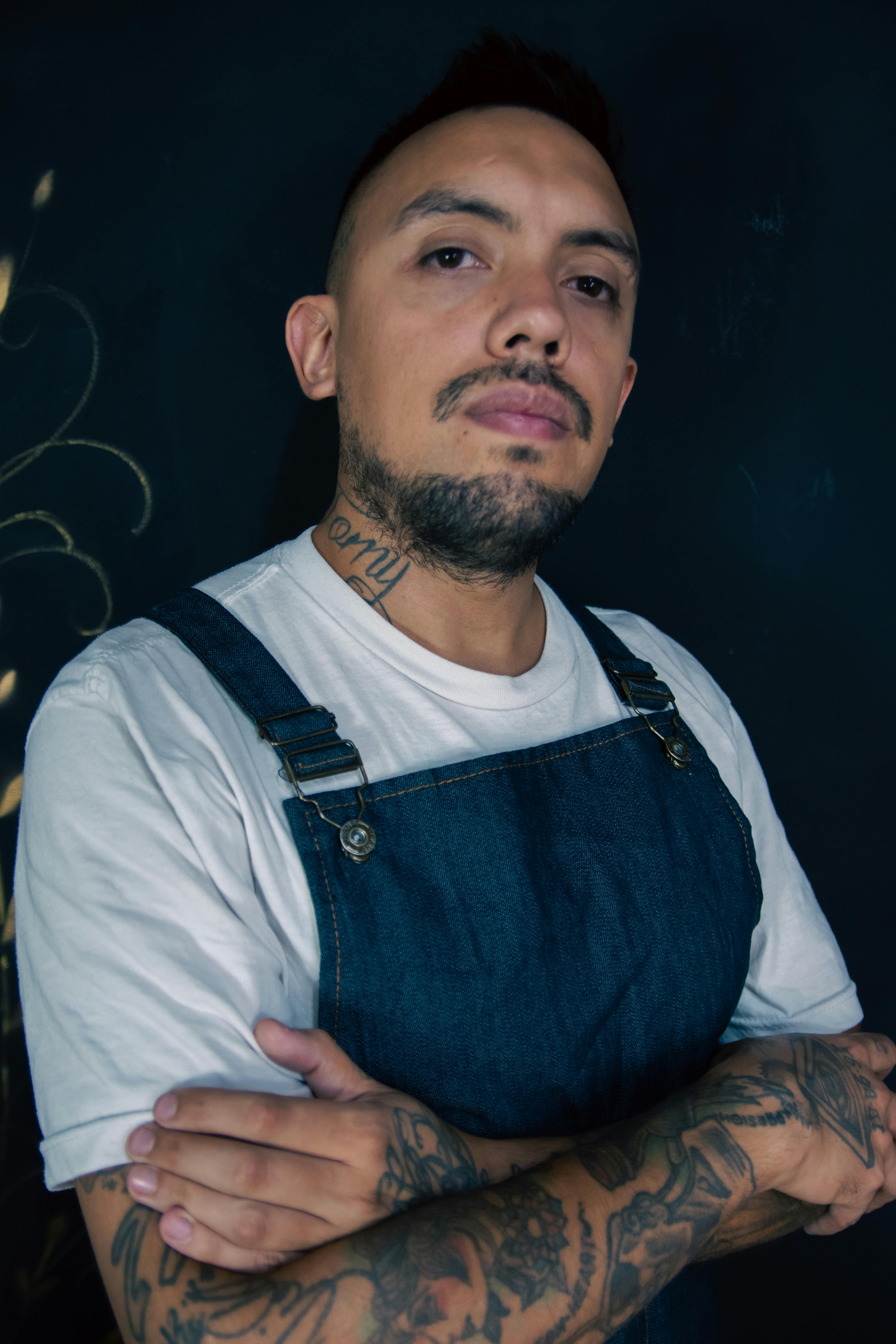 Tattooed man with beard and mustache, arms crossed wearing denim dungarees and white t-shirt.