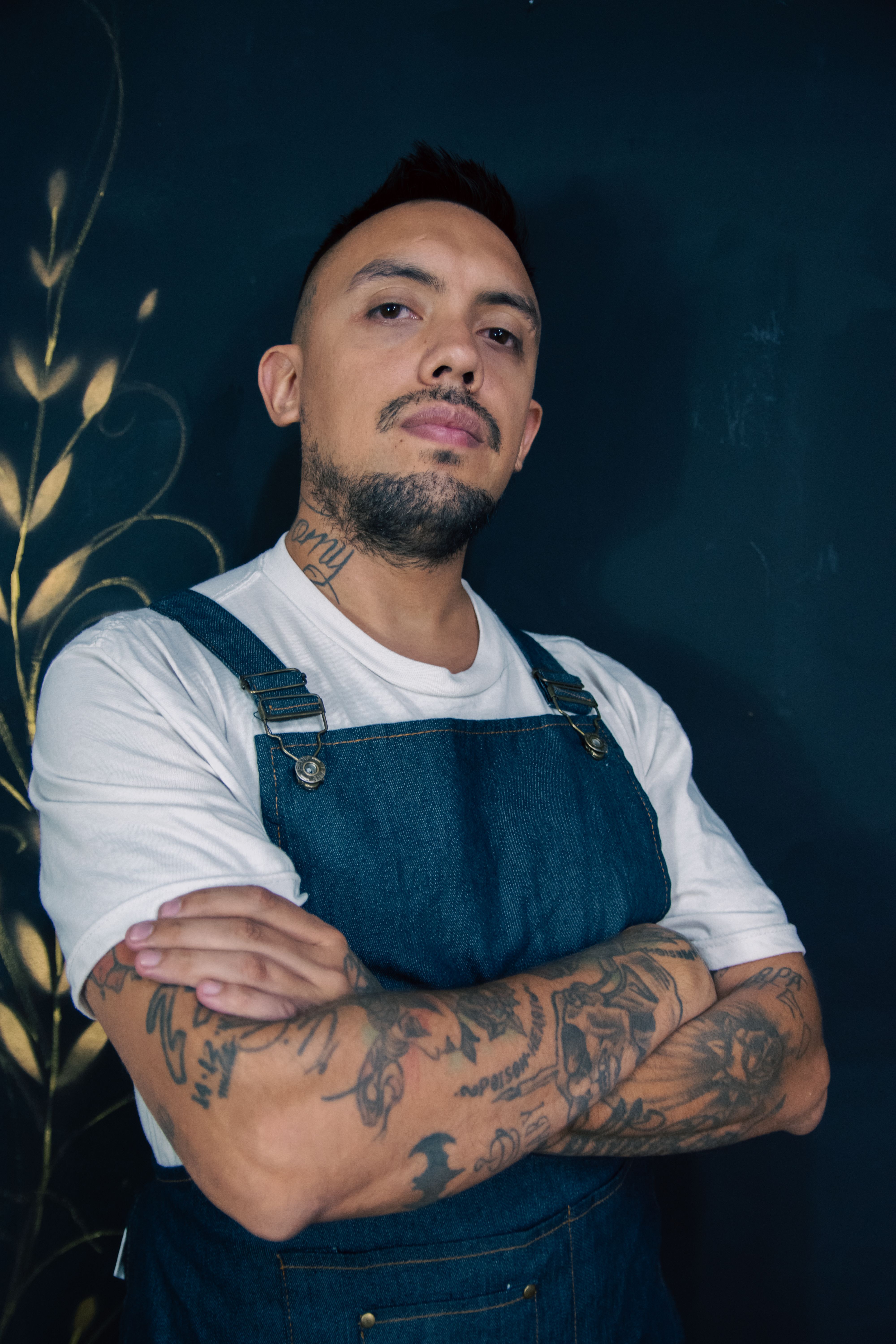 Confident tattooed man with crossed arms wearing denim overalls and a white t-shirt.