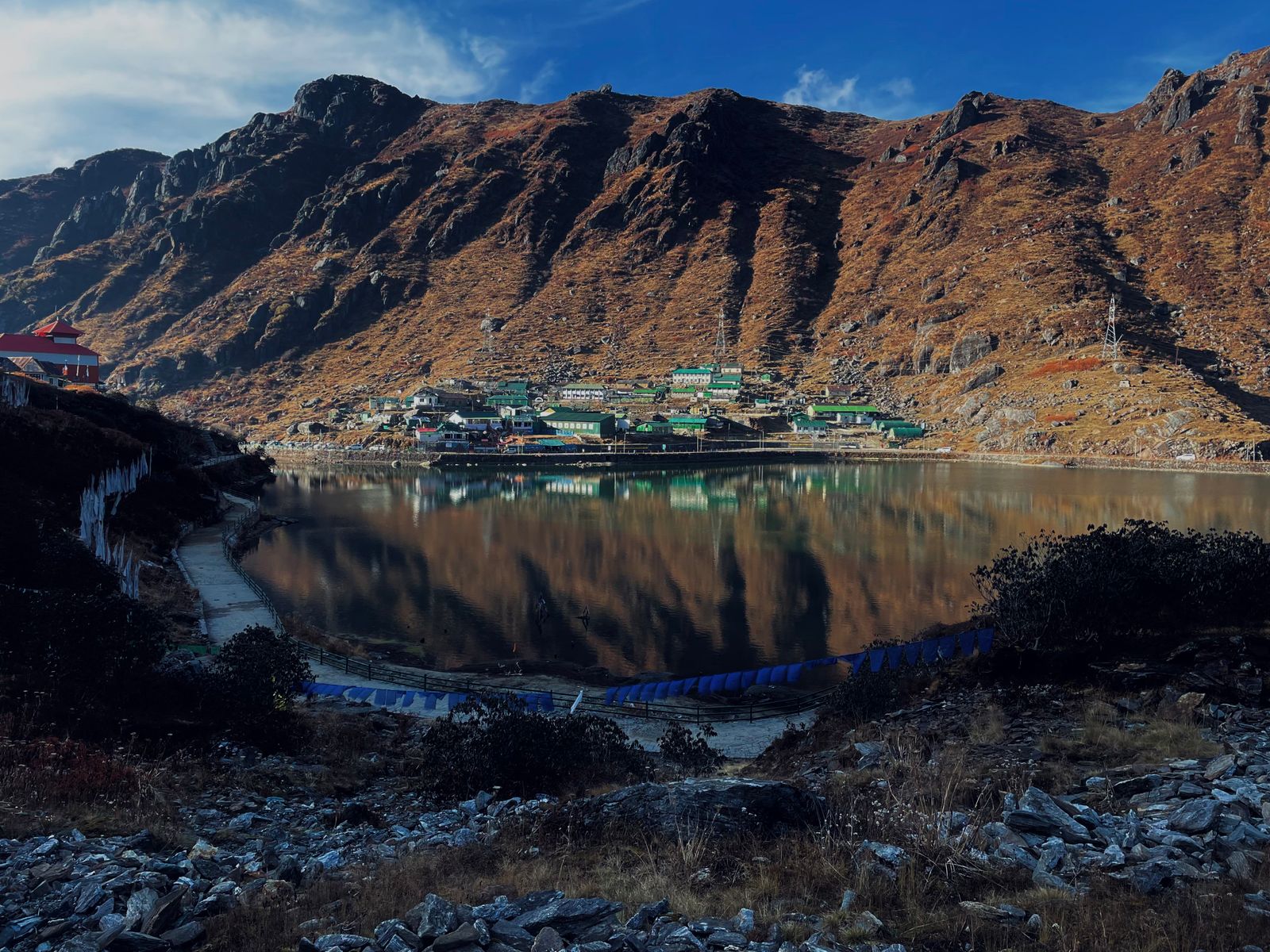 Tsomgo Lake still as glass under bare brown slopes, the green roofed lakeside settlement reflected in the water