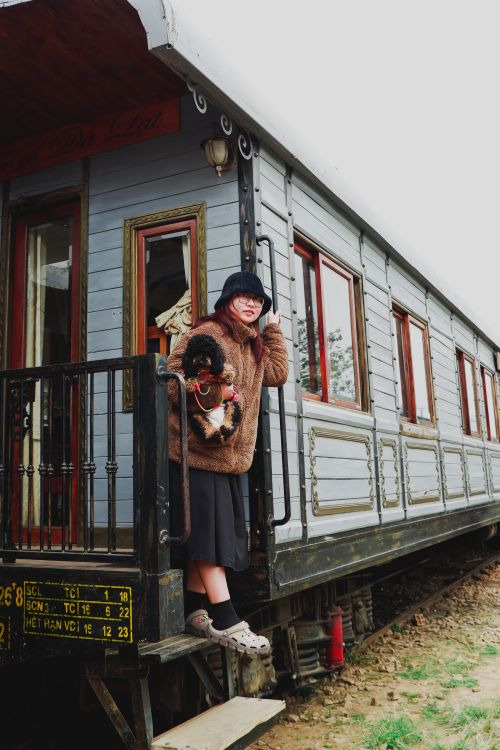 Free Woman in cozy attire holding a dog on a vintage train platform outdoors, showcasing travel mood. Stock Photo