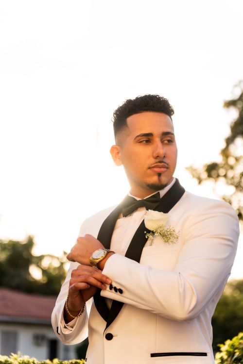 Free Bridegroom adjusting cuff, wearing a white tuxedo with boutonniere, backlit by sunset. Stock Photo
